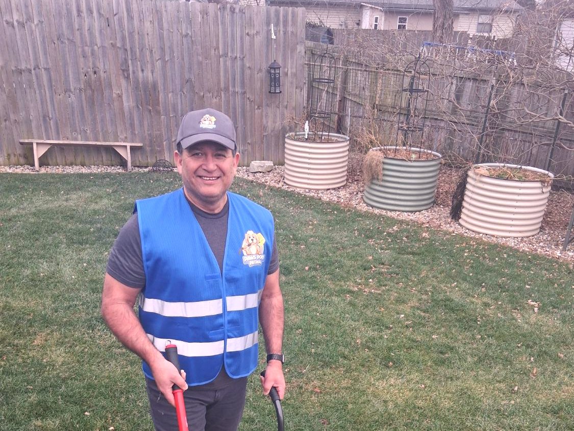 Charles Macias, owner of Paws Poo Patrol, smiling in a branded blue vest and cap in a fenced back yard